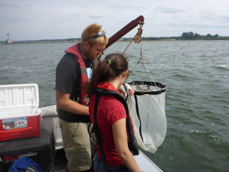 Two people stand on the back of a boat with a net on an arm with a winch