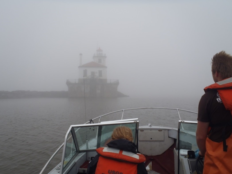 two people on a boat. It's foggy and one can barely see a lighthouse at the end of a breakwall near the boat.