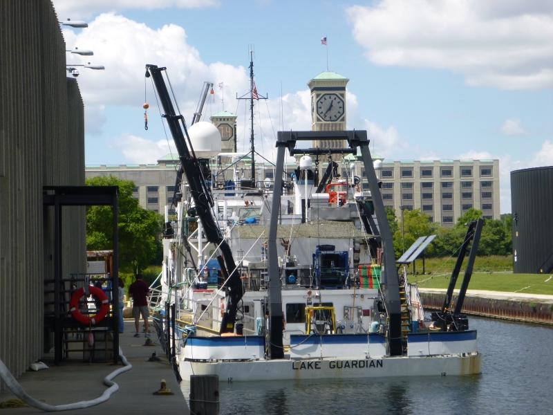 The stern of a large boat tied up at dock by a building with a clock tower. The boat is labeled "Lake Guardian"