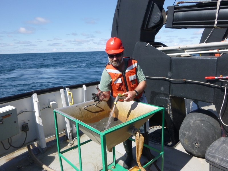 A person in a hat and life jacket stands by a tub of muddy water, holding up one muddy hand while using a hose with the other. He is on a large boat.