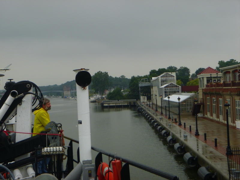 A person in a rain coat watches from the deck of a boat as it pulls up alongside a dock near a commercial building