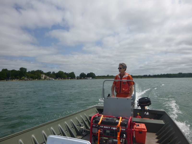 A person with graying hair and sunglasses stands at the console of a flat-bottomed boat motoring through a river