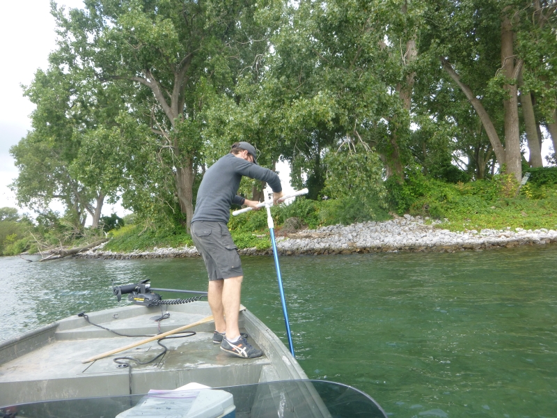 A person standing on the front of a boat. They are holding a long rod with a T on the end and using it in the water.