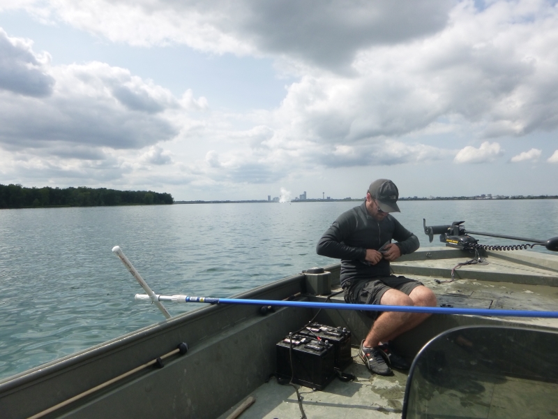 A person sitting in the front of a boat closing a plastic bag. There is a pole balanced across the boat in front of them, and a plume of mist in the distance behind them.