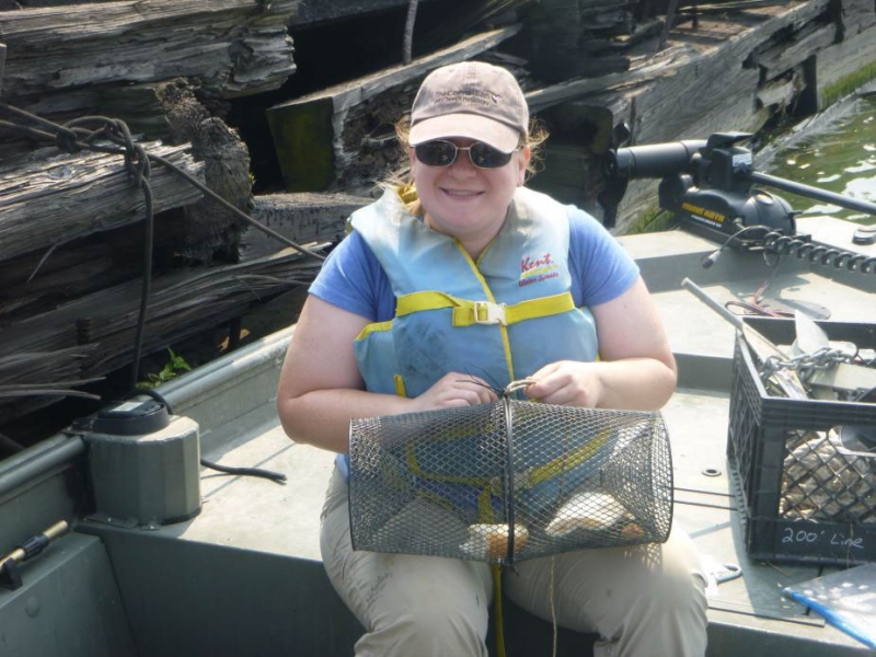 A person sitting on a small boat near a wall. She has a cylindrical mesh cage on her lap.