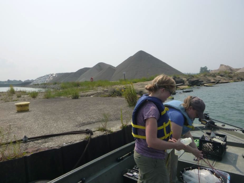 Two people on a boat near the break wall at a port. They are holding a bottle and a net. There are mounds of gravel in the background.