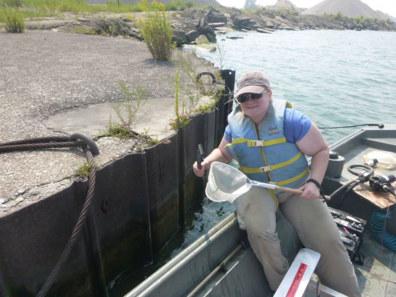 A person sitting on the edge of a boat near the break wall at a port. They are holding a net and a scraper.