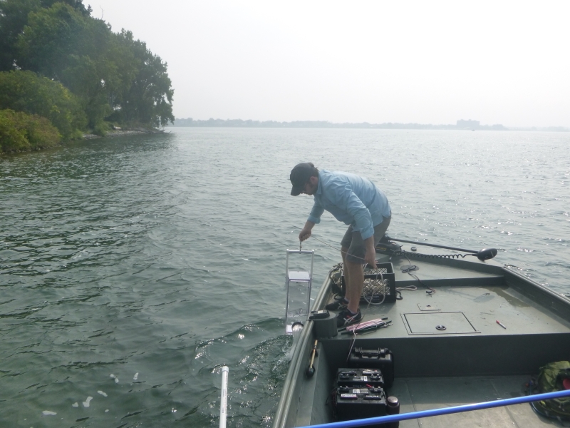 A person at the front of a boat pulls a clear box sampler out of the water.