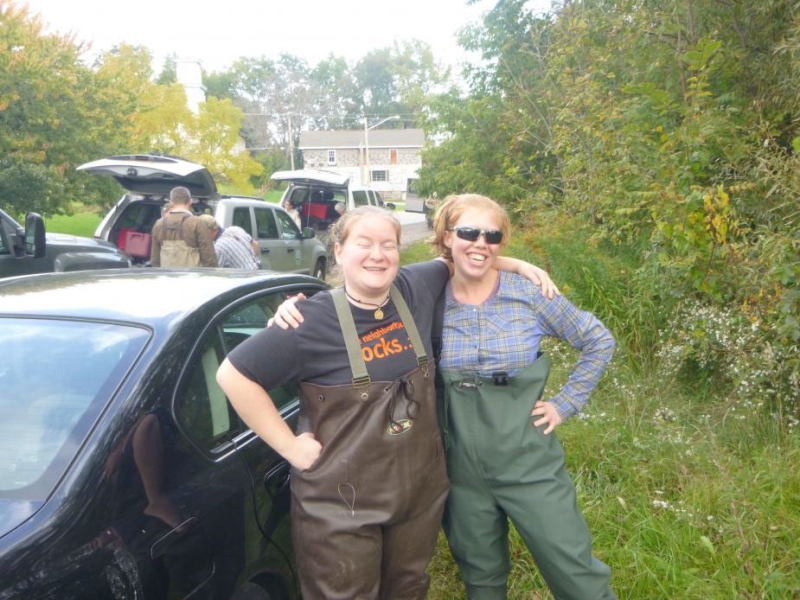 Two people wearing chest waders pose together next to a car. Trees and a few cars and people are in the background.