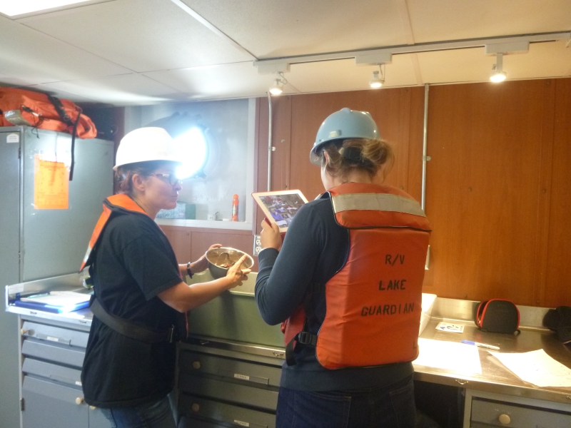 A person holds up a bowl of mud while another person points a tablet at it to record an image or video. They are both wearing hard hats and life jackets inside a lab on a boat.