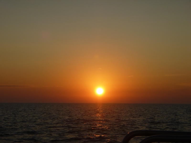 Sunrise over water. The sky is cloudless but hazy, intense red by the horizon and yellow for much of the sky above that. The sun is bright yellow.