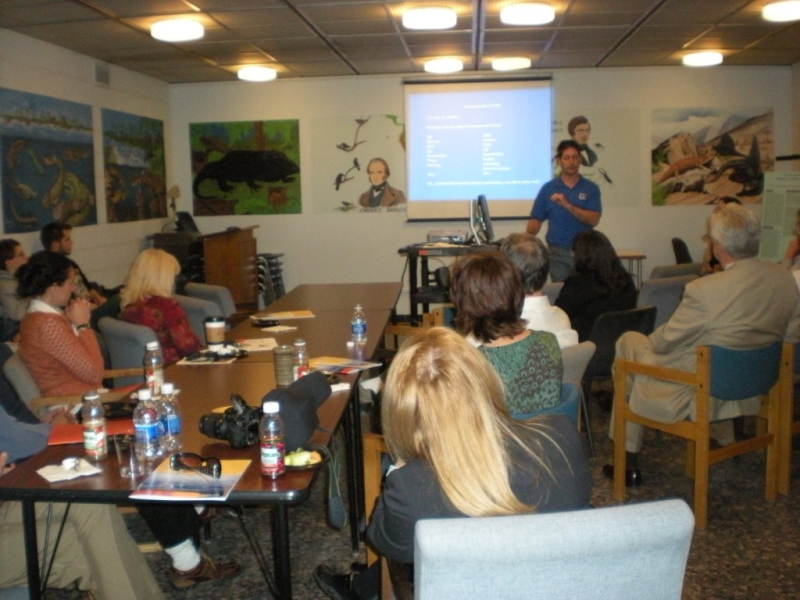 a group of people in chairs watch a presentation given by a different person at the front of the room. There is a presentation projected onto a screen, and murals painted on the walls of the room.