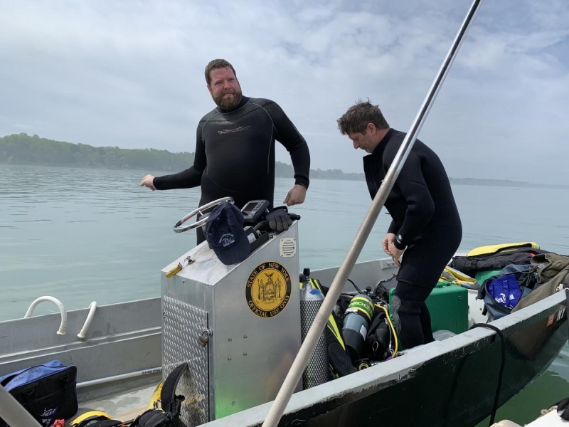 Two people in wetsuits stand in a boat on a body of water. There are air tanks next to them.