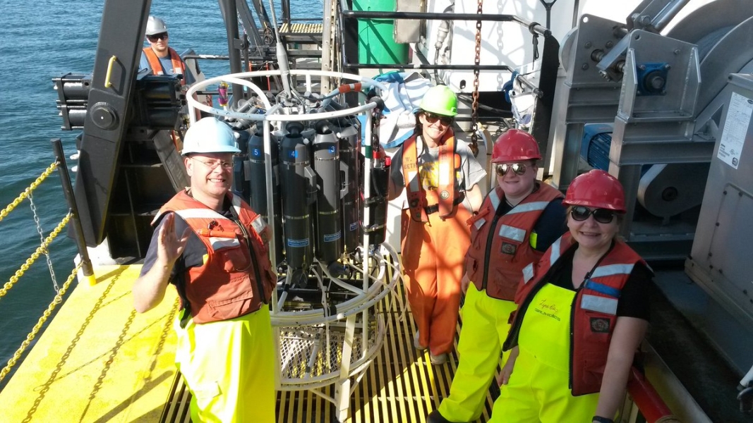 Four people stand on the deck of a large boat next to an instrument made of cylindrical bottles in a frame