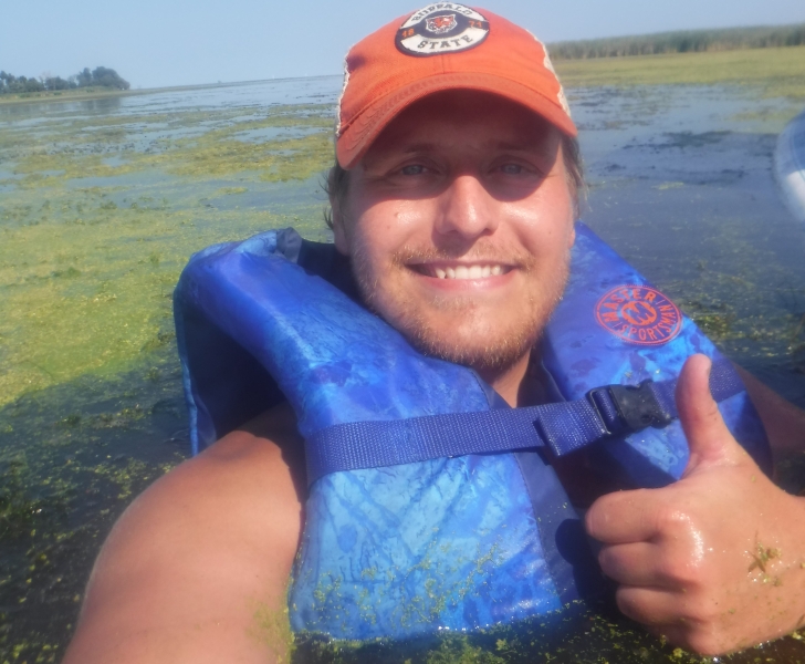 A person wearing a life jacket stands in water up to their chest as they take a smiling selfie with thumbs up.