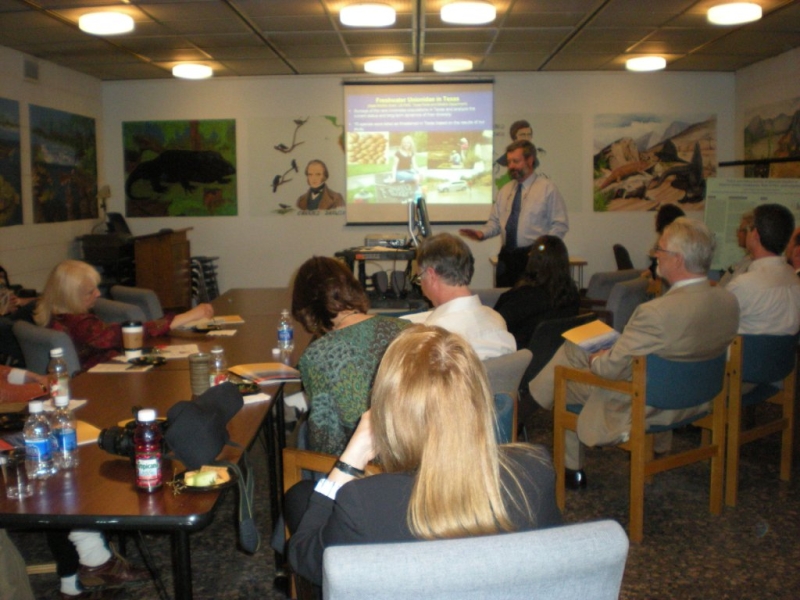 a group of people in chairs watch a presentation given by a person at the front of the room. There is a presentation projected onto a screen, and murals painted on the walls of the room.