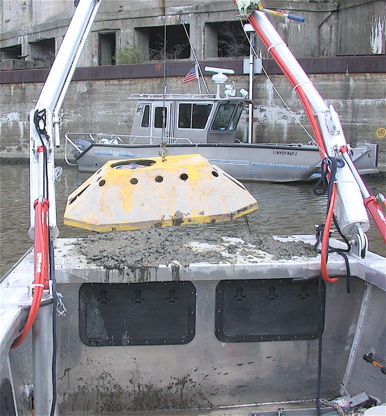 The frame on the front of a boat is used to lift a large metal device. There is mud on the boat. The boat is facing another boat labeled "Buffalo Police."