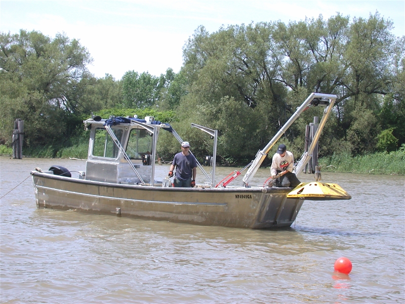 Two people on a boat. The frame on the front of the boat is lifting a large metal device.