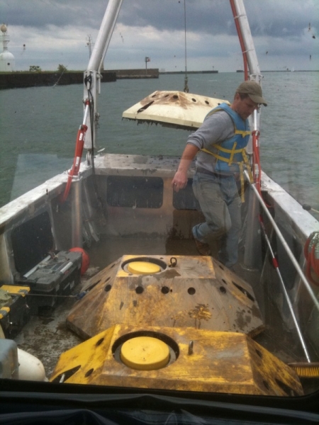 A person in the front of a boat. There are two large metal devices on the deck and the frame at the front of the boat is lifting a third one.