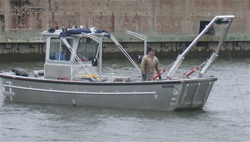 One person works in the front of a boat with an A-frame on the front of it. Another person is in the cabin.