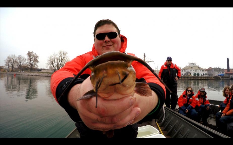 A student on a boat holds up a brown bullhead to the camera. There are students in a second boat in the background.