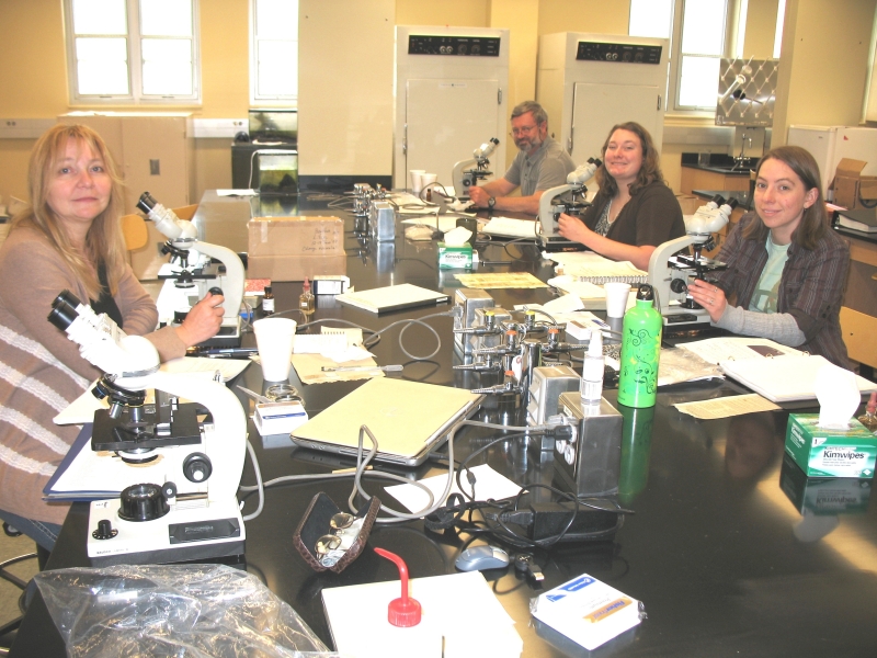 Four people sit at microscopes on either side of a large lab table