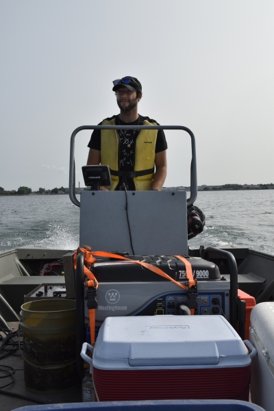 A graduate student wearing a life jacket stands at the steering console of a small boat in a river.