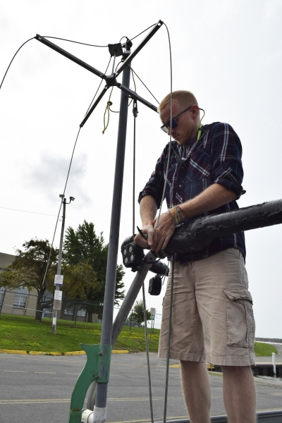 A person stands at a railing tying a rope on a boat in a parking lot. The rope holds a pole with wires in a vertical position.