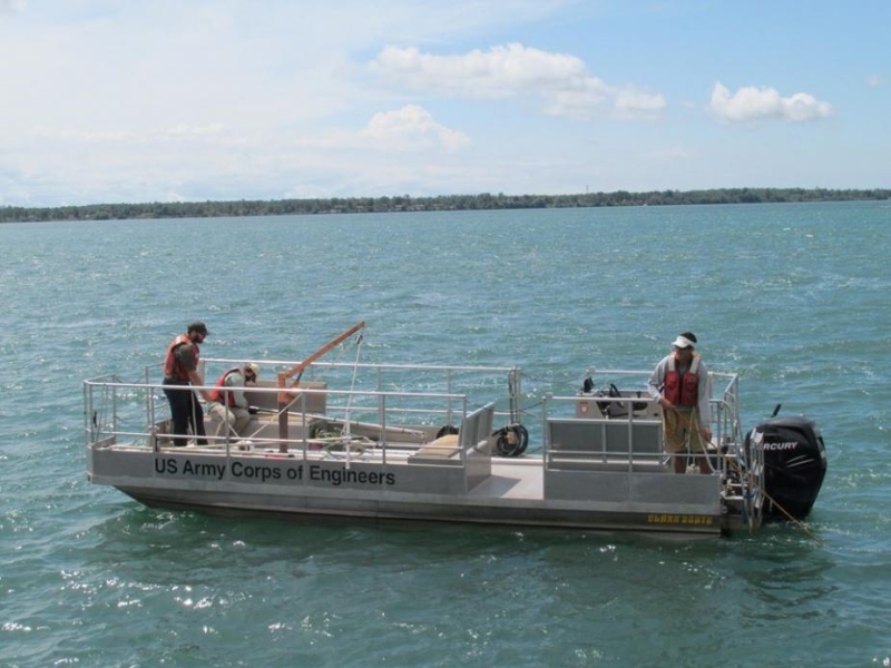 Three people on a small boat with railings labeled "US Army Corps of Engineers."