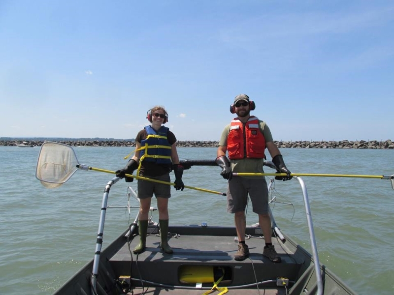 Two people stand in the front of a boat wearing life jackets, ear protection, rubber gloves and rubber boots, and holding nets on long poles.