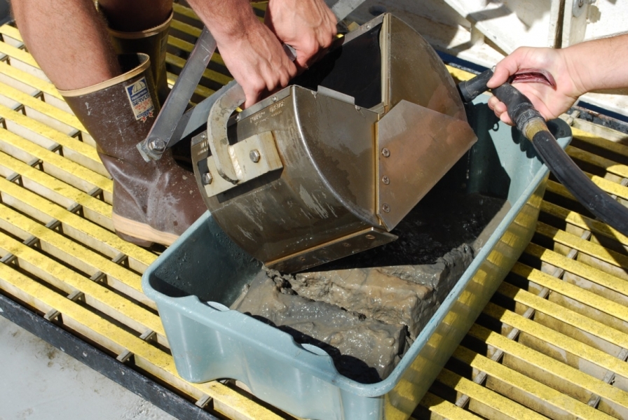 Someone opens a metal grab sampler into a tub and while a second person rinses the mud inside into the tub