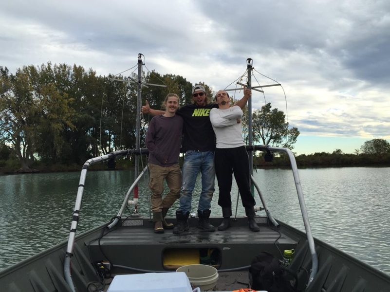 Three men pose for a picture at the front of a boat. The one in the center has his arms around the others with thumbs up, and the one on the right has arms crossed with a confident stance