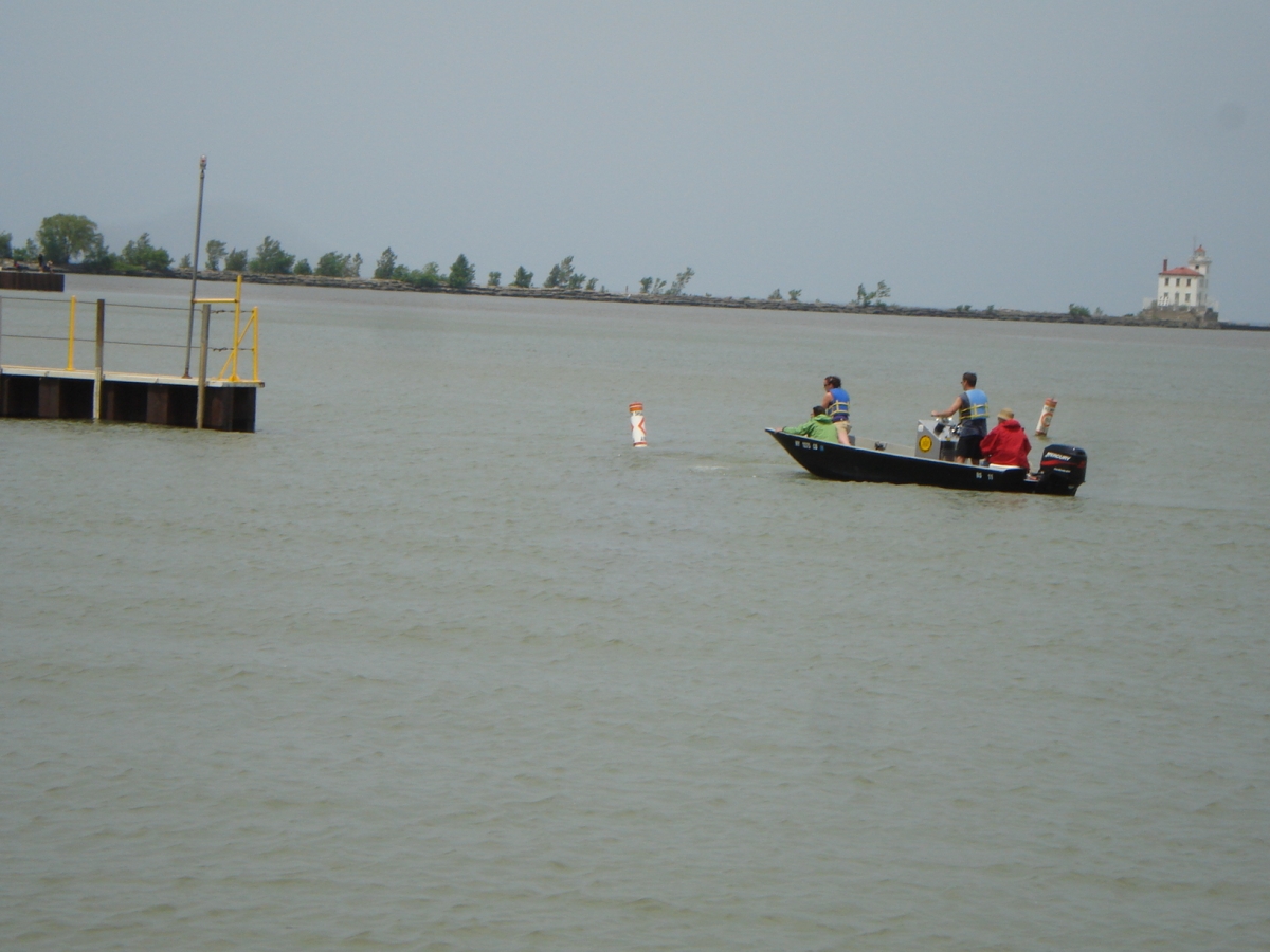 A small boat with a few people on it near a dock. There is a lighthouse in the distance.