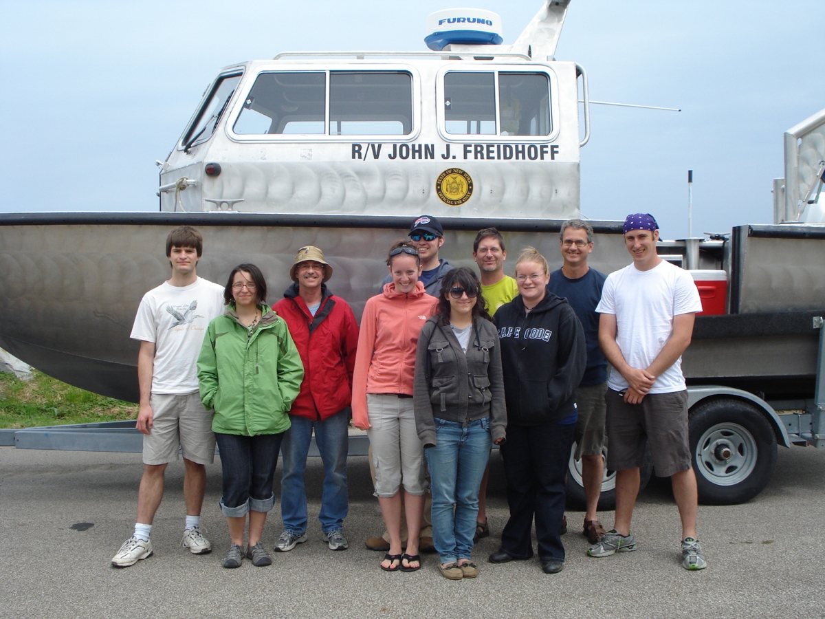 Ten people stand in front of a boat on a trailer. The boat is named 