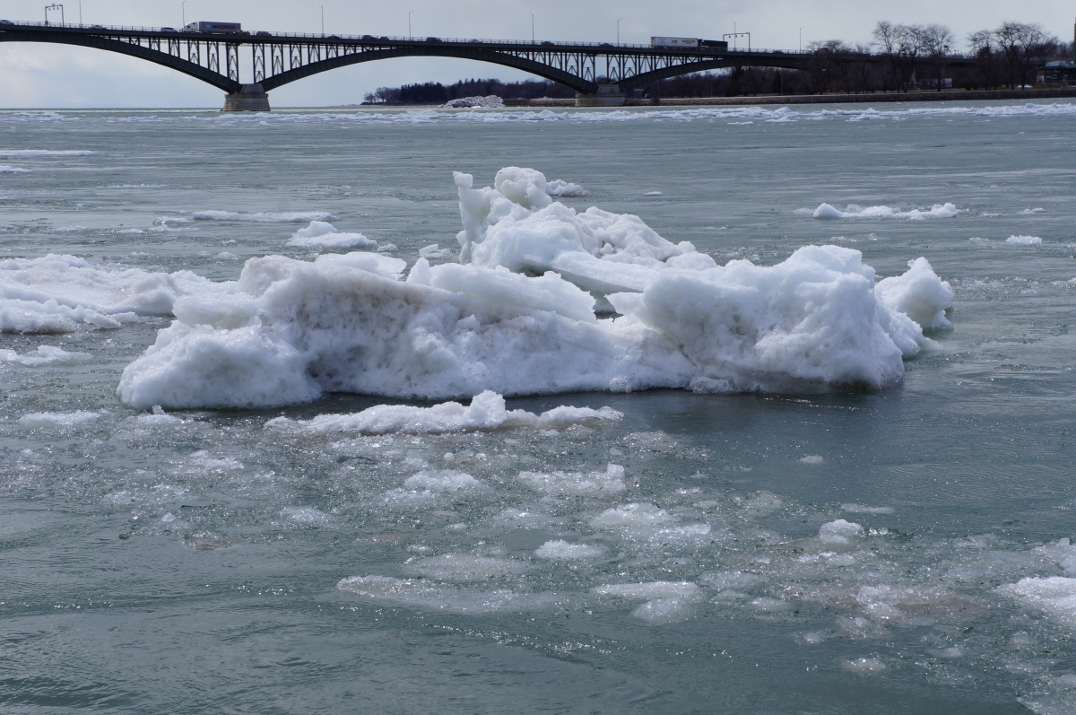 A large chunk of ice in an icy river. There is a bridge in the distance.