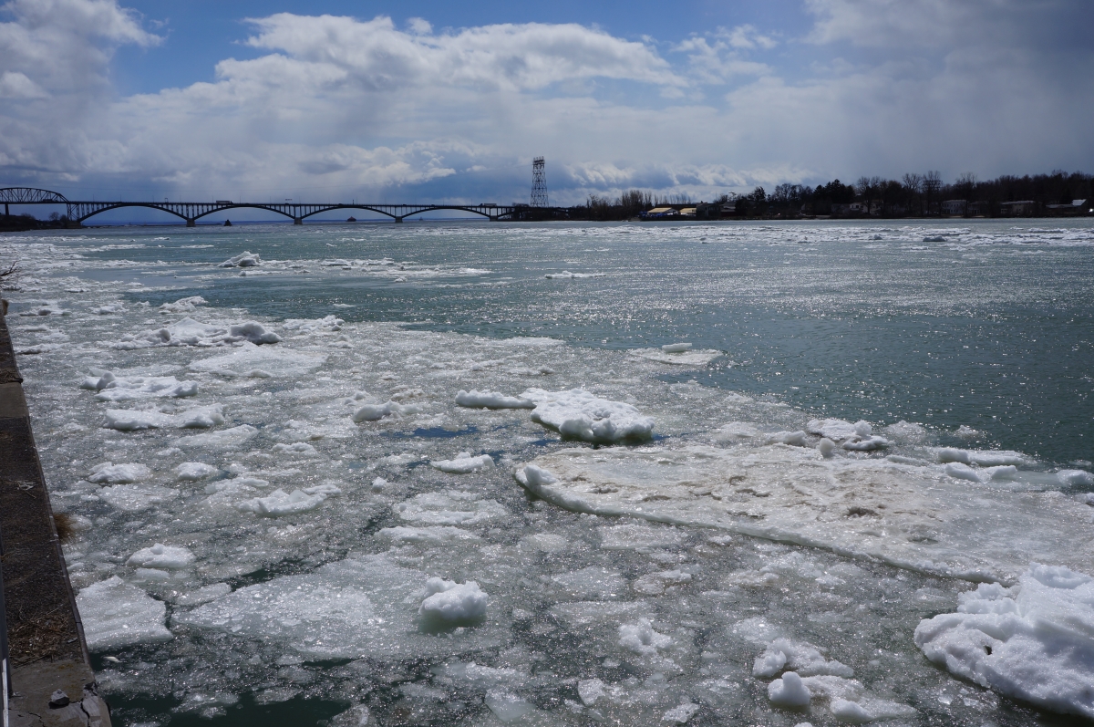 Large chunks of ice in the river. There is a bridge in the distance.