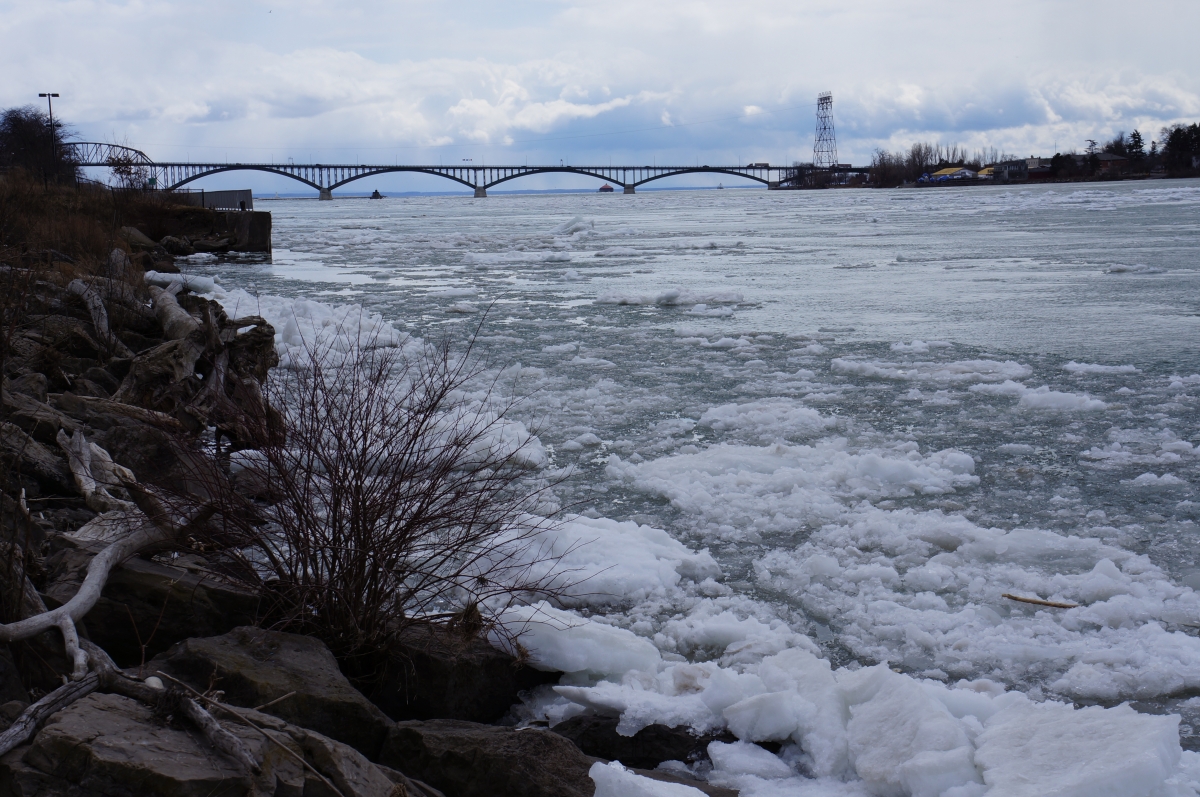 Ice piling up along the shore of the river. There is a bridge in the distance.