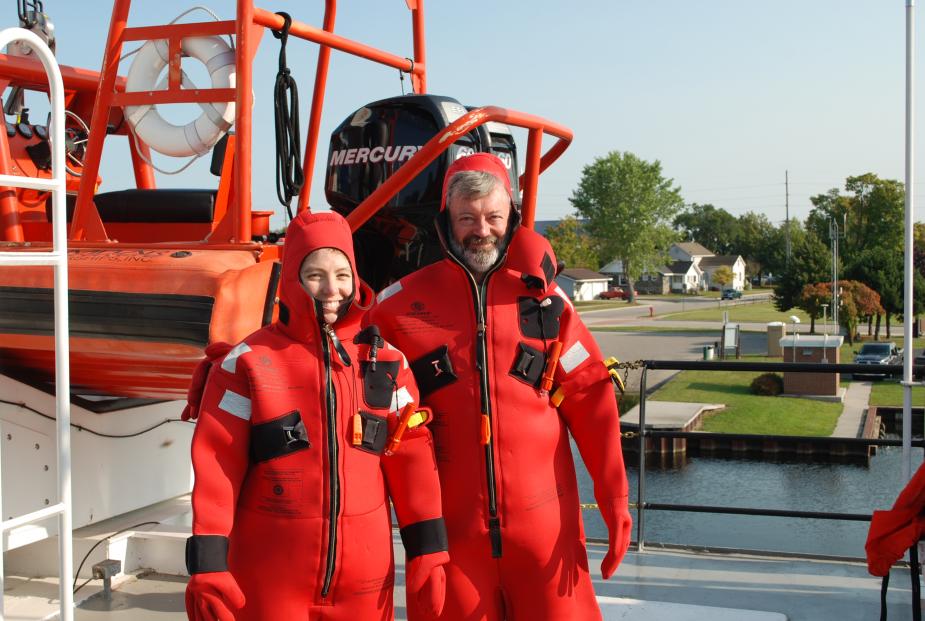 Two people wearing red survival suits on the deck of a large boat. There is an emergency rescue watercraft on deck behind them.