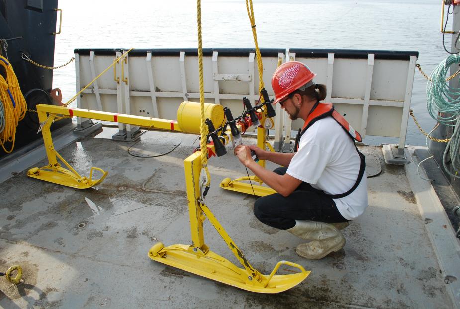 A person kneels before a metal frame with sled feet on the deck of a large boat. They are turning on lights and adjusting a camera.
