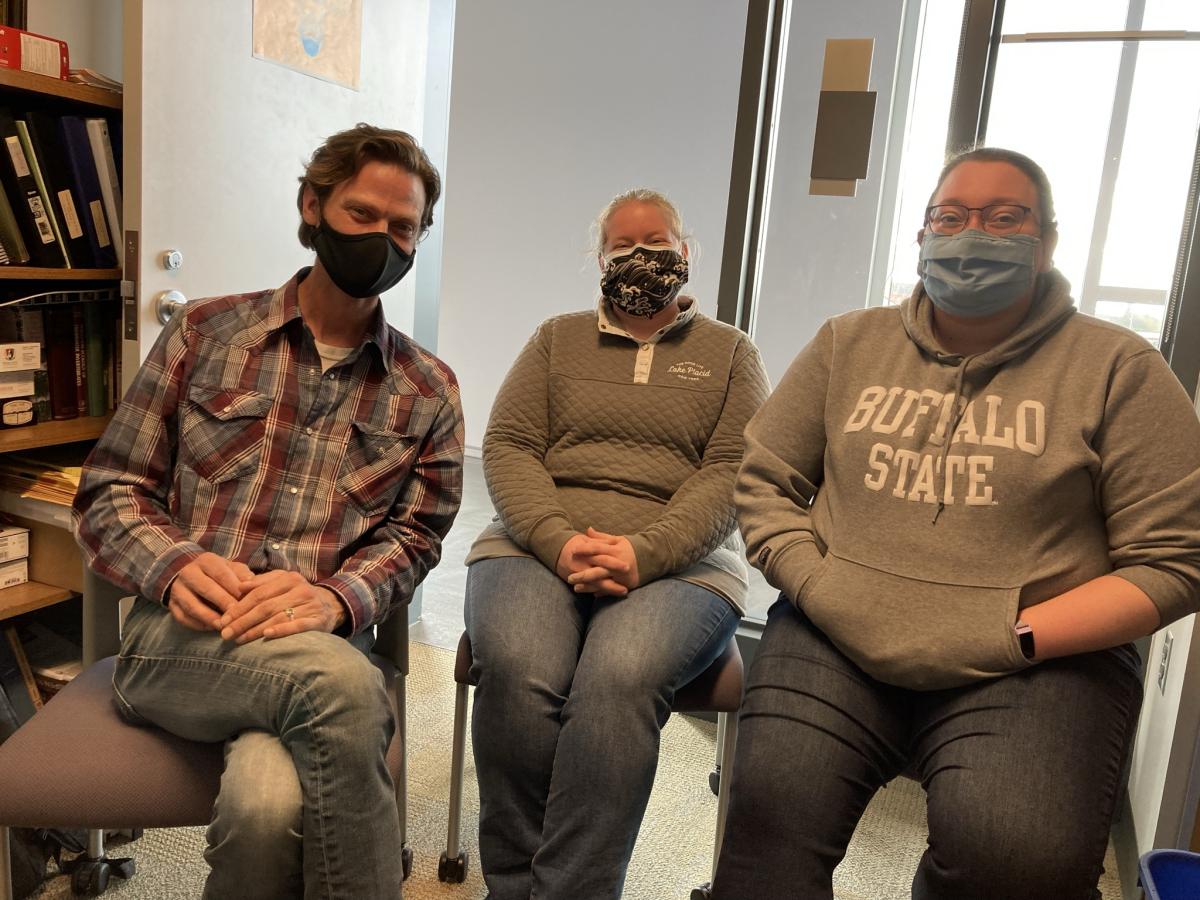 Three smiling people wearing face masks sit in chairs next to each other in a small office. There is an open door behind them.