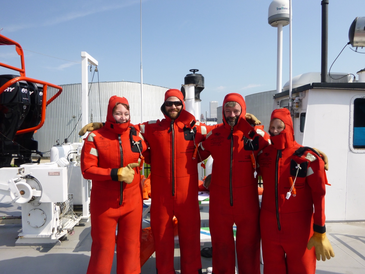 Four people in red survival suits on the deck of a large boat