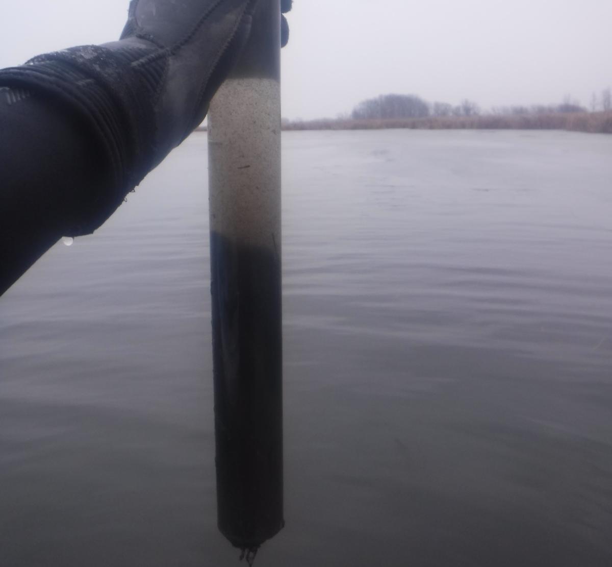 A gloved hand holding up a plastic tube with water and sediment in it. In the background is a calm body of water on an overcast day.