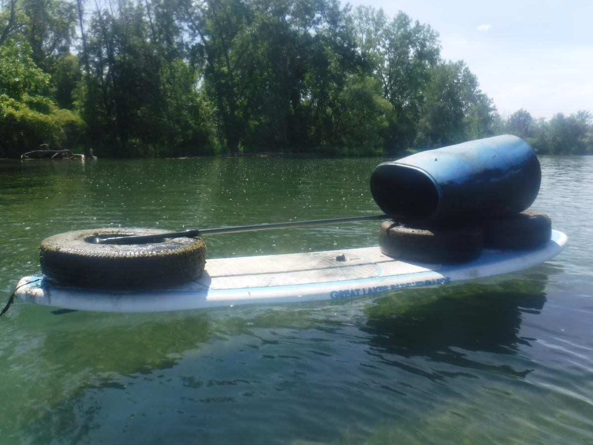 A paddleboard floating on a body of water near the shore with three tires and a plastic barrel on it. The paddle is laid over the tires.