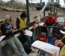 Students sit on a boat tied up at dock while a person in a floatation coat holds up a research instrument