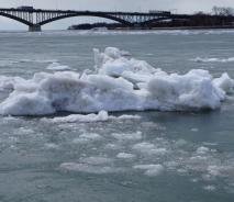 A large chunk of ice in an icy river. There is a bridge in the distance.