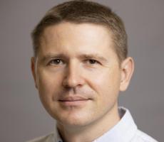 A light-skinned man with short brown hair wearing a light blue striped collared shirt.