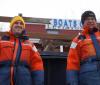 two people in orange and blue float suits and knit caps stand in front of a building with a sign "Boats"