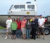 Ten people stand in front of a boat on a trailer. The boat is named "R/V John J. Freidhoff"