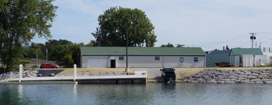 A white building with a green roof with a dock and boat launch, next to the water. A boat is backed into the water with a trailer.