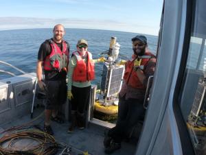 Three people smile on the back of a boat holding a squat buoy with solar panels and weather instruments.